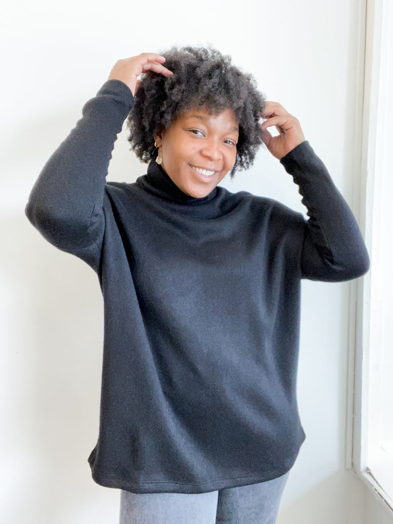 This is a nearly full length picture of a model. She is wearing a light long sleeve turtle neck sweater in black.